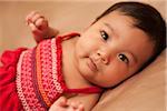 Close-up portrait of Asian baby lying on back, wearing red dress, looking at camera, studio shot on brown background