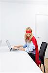 Portrait of happy businesswoman in superhero costume using laptop at office