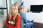 Annoyed couple in Christmas sweaters and party hats sitting back to back at home