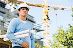 Male architect holding rolled up blueprints while standing at construction site