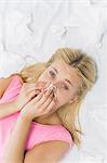 High angle portrait of young woman blowing nose in tissue paper while lying on bed