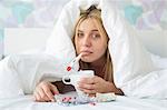 Portrait of sad woman with coffee mug taking temperature while wrapped in quilt on bed