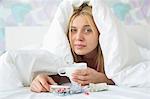 Portrait of young woman with coffee mug and medicines suffering from fever while covered in quilt on bed