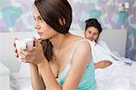 Worried young woman holding coffee mug while ill man relaxing in background