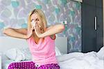 Young woman blowing her nose in tissue paper while sitting on bed