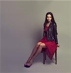 Sexy Brunette Woman in Red Fashion Dress and Leather Jacket Sitting on a Chair. Copy Space. Toned Photo.