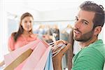 Bored man sitting with shopping bags while woman by clothes rack in the background