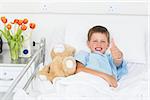 Portrait of little boy gesturing thumbs up with teddy bear in hospital bed