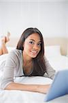 Pretty girl lying on bed using her tablet smiling at camera in her bedroom at home