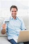 Smiling man using laptop sitting on sofa shopping online at home in the living room