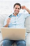 Cheerful man using laptop sitting on sofa shopping online at home in the living room