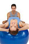 Male trainer helping young woman stretch on fitness ball over white background