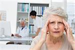 Close-up of a senior patient suffering from headache with doctors in the background at the medical office
