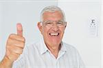 Portrait of a smiling senior man gesturing thumbs up with eye chart in the background at medical office