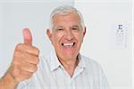 Portrait of a smiling senior man gesturing thumbs up with eye chart in the background at medical office