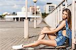 beautiful girl with long legs in jeans short overalls sits on skateboard near fence