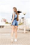 beautiful smiling girl with long legs in jeans short overalls stands with skateboard on shoulder