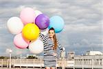 beautiful young lady in black and white striped short dress holds a bunch of multicolored balloons smiling for camera