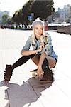 Blond girl in short shorts, jeans coat, black stockings and grey hat sits on skateboard and bites sunglasses