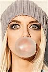 Portrait of beautiful young blond girl in beanie hat blows big bubble from bubble gum