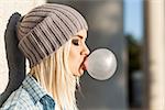 Portrait of beautiful young blonde teenage girl in beanie hat blows big bubble from bubble gum