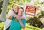 Happy Couple Holding House Keys In Front of Sold Real Estate Sign.