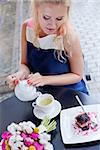 a beautiful young blond girl in summer dress at the table in pavement cafe is pouring green tea from the teapot into a white ceramic cup