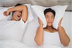 Annoyed woman covering her ears with pillows to block out snoring at home in bedroom