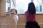 Girl and her pet dog standing and looking out of window