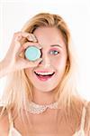 Close-up portrait of young woman holding blue macaron up to her eye, looking at camera and smiling, studio shot on white background