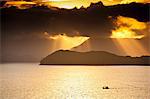 South America, Brazil, Green Coast (Costa Verde), Sao Paulo, Ubatuba, a fishing boat sails in front of rainforest-clad mountain spurs, both silhoutted against a golden sunset