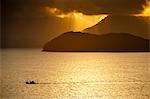South America, Brazil, Green Coast (Costa Verde), Sao Paulo, Ubatuba, a fishing boat sails in front of rainforest-clad mountain spurs, both silhoutted against a golden sunset