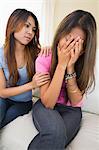 Tearful woman sitting on a white couch being consoled by her sister at home in living room