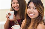 Two pretty brunette sisters having a cup of coffee while sitting on a couch and looking at the camera