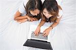 Two young sisters lying on a bed using laptop at home in bedroom