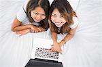 Two happy sisters lying on a bed using laptop smiling at the camera