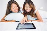 Two smiling brunette sisters using a tablet while lying on a bed