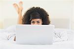 Pretty smiling  brunette lying on bed hidden behind laptop in bright bedroom