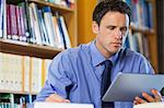 Frowning librarian sitting at desk using tablet in library in a college