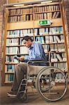 Frowning man sitting in wheelchair reading a book in library in a college