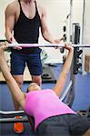 Trainer helping lying toned woman exercising with barbell in weights room of gym