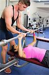 Handsome instructor helping lying woman exercising with barbell in weights room of gym