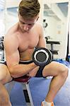 Muscular topless man sitting on bench training with dumbbells in weights room of gym