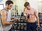 Instructor taking notes of man lifting dumbbells in weights room of gym