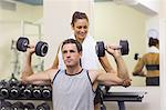 Trainer correcting muscular man lifting dumbbells in weights room of gym