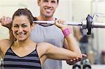 Instructor helping smiling woman lifting barbell in weights room of gym