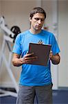 Serious personal trainer with clipboard standing in the gym