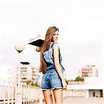 beautiful smiling girl with long legs in jeans short overalls stands with skateboard on shoulder