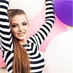 close-up of beautiful smiling girl with smokey eye make up, ponytail hair in black and white striped dress keeps her hands raised holding bunch of multicolored balloons