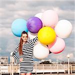 beautiful young lady in black and white striped short dress holds a bunch of multicolored balloons smiling for camera